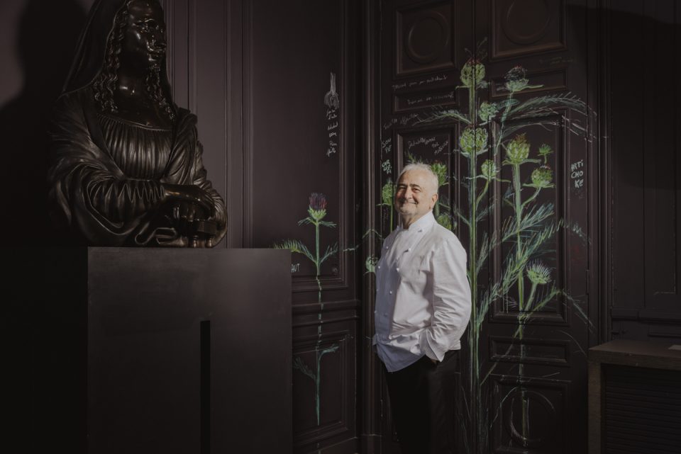 Le restaurateur Guy Savoy dans son restaurant a la monnaie de Paris vue de trois quart devant un mur type hausslmanien gris fonce abec illustrations chardons et une sculpture en premier plan